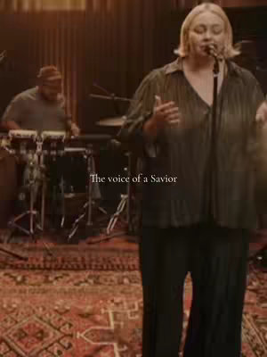 The image showcases a live performance of a female vocalist in a recording studio. The scene is warm-toned, with a large patterned rug filling the foreground. The vocalist is center-frame, singing into a microphone, and is wearing a dark, flowing top over dark pants. Visible behind her is a drummer and other band members in the background, adding to the music theme. Overlaid text says 'The voice of a Savior.'