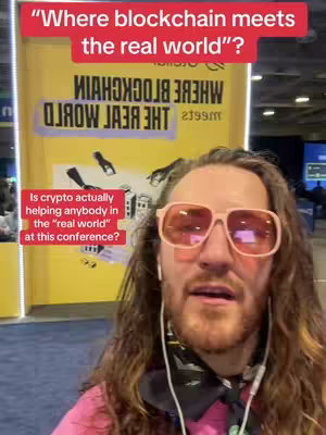 A man with long hair and pink-tinted glasses stands in front of a yellow banner with text that is partially visible, the man is wearing a pink shirt and white earphones. He is looking at the camera.