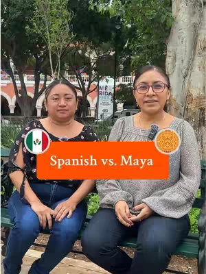 Two women sit side-by-side on a park bench with a tree and building background. An orange rectangle over the image states 'Spanish vs. Maya' with two circular elements around each woman; the left one shows a Mexican flag, and the right one has a Maya glyph. The women appear in front of green foliage with the building and some people walking around in the background.