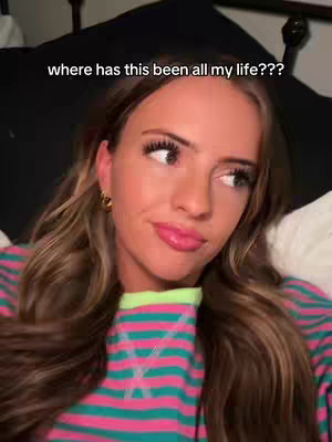 A woman with long, wavy brown hair is turned to the side, looking off-camera with a slightly amused expression. She's wearing a colorful striped shirt. The text overlay reads 'where has this been all my life???'. The background is blurred, with what looks like a dark bed and white pillows.
