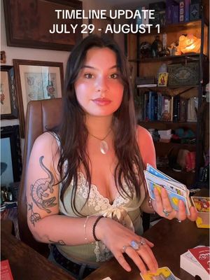 A young woman with long brown hair sits at a wooden table, holding a deck of tarot cards. She wears a light green top and has a tattoo on her left arm. Text at the top reads 'Timeline Update July 29 - August 1'. The background includes a bookshelf and framed pictures.
