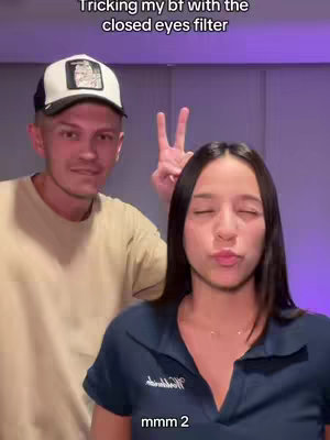 A couple in a selfie-style shot. The man is on the left, wearing a baseball cap, and smiling at the camera on the left The woman is in the center, with a closed-eyes filter on. The background is blurred and the color behind them is purple. A text overlay says 'Tricking my bf with the closed eyes filter' in the top left of the image.