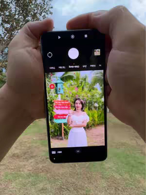 A person is holding a smartphone with both hands, showcasing the phone screen on which an image of a woman is displayed. The person appears to be outside because there is foliage in the background, including green grass and trees. The screen displays the woman posing with a sign behind her. The composition uses natural lighting and the primary colors visible include green, white, black, pink and red.