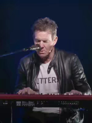 A man with short, graying hair wearing a black leather jacket over a white t-shirt is playing a red keyboard. He's singing into a microphone. The background is a dark, blurred setting which focuses attention on him.