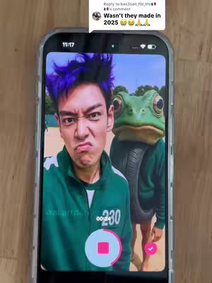 A selfie taken in front of a green screen, with a person wearing a green Squid Game jumpsuit making a disgusted face and another person wearing a frog costume, plus text overlayed on the phone screen.