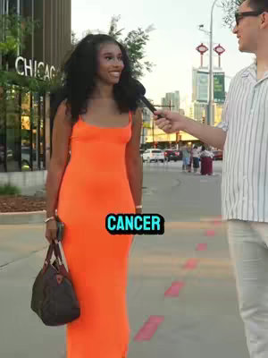 A woman in an orange dress is being interviewed. The background shows an urban setting with buildings and a sidewalk. Text that says 'CANCER' is overlaid on the image.