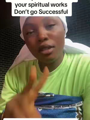 A woman with brown skin is centered in the frame, wearing a white head covering and a light green t-shirt. She is making a peace sign with two fingers. The background is a blurred neutral color.