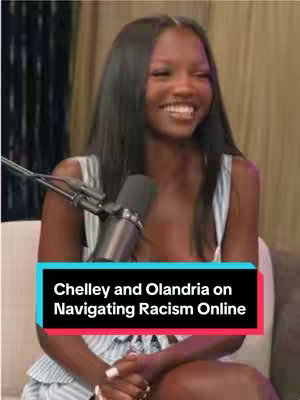 A woman with long, dark hair sits smiling in front of a microphone. Behind her, there is a blurry background that appears to be a curtain. The text overlay at the bottom of the screen reads 'Chelley and Olandria on Navigating Racism Online', set against a black and teal background.