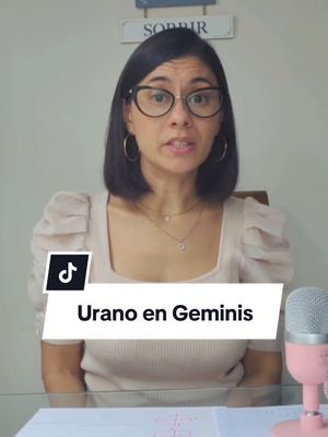 A woman with short dark hair and glasses is looking directly at the camera and speaking. She is wearing a light-colored top. Below her is a white banner with the text "Urano en Geminis". In the background is a light-colored wall and a microphone.