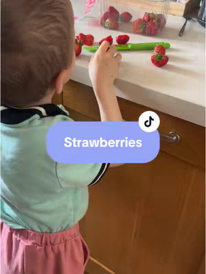 A slightly over-the-shoulder shot shows a young child from the neck down, standing in front of a countertop, reaching for strawberries. The child is wearing a light blue shirt with black and white trim, and dark pink pants. The hook is a blue oval displaying the word 'Strawberries'. A white TikTok logo is in the top right corner of the hook, with the TikTok logo visible. Strawberries and a green knife are also present on the counter.