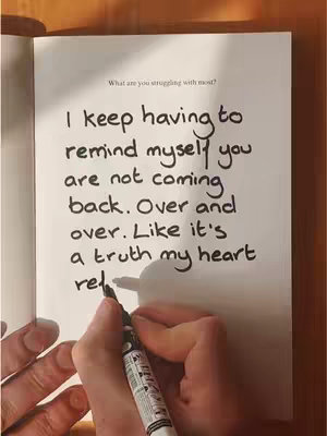 The image shows a book open to a page with handwritten text. A hand holding a pen writes on the page. The text is in black ink on white paper.