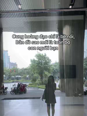 The image presents a wide exterior shot with a modern architectural style. Tall, vertical glass panels create a repeating linear pattern across the facade. In the foreground, a person stands, slightly obscured by the shot, revealing a background with motorcycles and cars in motion. The sky is visible above the building, suggesting an outdoor setting. The image uses a slightly muted color palette, giving a contemporary feel, with the primary focus on the contrast between the building's lines and the softer, natural elements of the sky and trees through the entrance.