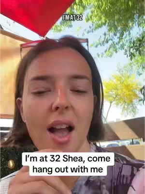 A woman is seen in a selfie-style shot with a blurred outdoor background. The upper part of her torso and face are in focus. A red canopy is seen in the background and green foliage. There is text overlaying the top and bottom of the image.