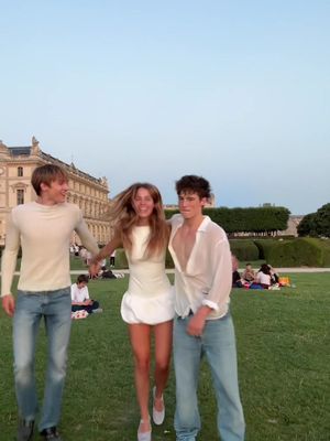 Three young adults are captured in a bright, outdoor setting. The focus is on a woman in a white dress who is running forward, holding hands with two men, one on each side. The background is blurred, suggesting a park or public garden lit in the evening.