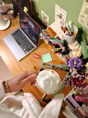 A well-lit, overhead shot captures a study setup with a laptop, various stationery items, and a cup of what appears to be cream or foam. The composition includes a laptop with a visible screen showing an editing program, a cup of what appears to be a latte, pens, markers etc on a lightly colored wooden desk, with a hand holding the cup in the foreground, and a fluffy pink blanket partially visible, and some flowers on the right side. The color palette is warm and inviting, with a focus on the desk as the central element.