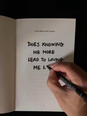 A hand holding a black pen writes on an open book. The left page is blank while the right contains the text: "DOES KNOWING ME MORE LEAD TO LOVING ME L". The background is dark except for the book. The lighting is soft and natural.