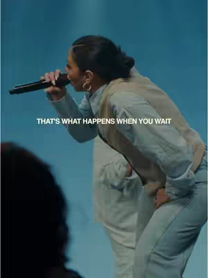 A woman with a microphone is singing on stage. Her back is turned to offer a profile of her face and a portion of her side. The background is a gradient blue. The text overlay reads, "THAT'S WHAT HAPPENS WHEN YOU WAIT".