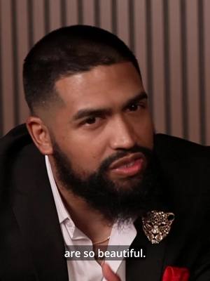 A man with a dark beard and short hair is the focus, seen from the chest up. He wears a black suit jacket over a white collared shirt. A gold necklace and a jeweled pin are visible. The background is a light, neutral color, with lines. The man appears to be speaking.