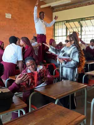 A group of people in a classroom setting, some wearing maroon and white uniforms, are interacting. A person on top of a desk raises their arms in celebration, while a woman in sunglasses holds a magazine near a desk where a person is reading it, with the title clearly displayed. The room has wooden desks and a light-colored brick wall.