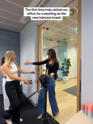 A woman in the foreground is entering an office space with arms open, and a woman standing in the doorway also with arms open. Text on the image reads, 'The first time Indy visited our office for a meeting on the new haircare brand,' and the office appears to be a modern corporate setting.