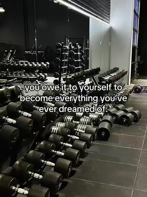 The image shows a well-lit gym with rows of dumbbells in the foreground. The weights are dark against the light-colored industrial floor. In the background, there are more weight racks and a large mirror. The overall tone is motivational, hinting at fitness and self-improvement.