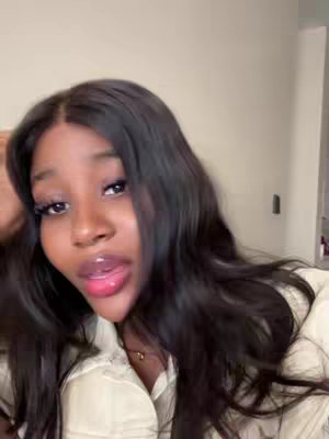 A selfie with a young Black woman at the center, looking at the camera with her mouth slightly open. She has long black hair. The background is a plain wall. She's wearing a light-colored top.