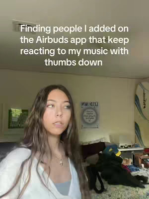 A young woman stands in a room, looking towards the central camera. Behind her is a bedroom with unmade bed with a colorful comforter and decorations scattered around. Above her head, white text reads "Finding people I added on the Airbuds app that keep reacting to my music with thumbs down."