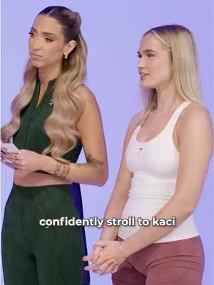 Two women are standing side by side against a light purple backdrop. The woman on the right is wearing a white tank top and red shorts, with her hands clasped in front of her. The woman on the left is wearing a dark green outfit and a serious expression, holding a paper in her hands. The image is well-lit, with even lighting across the subjects.