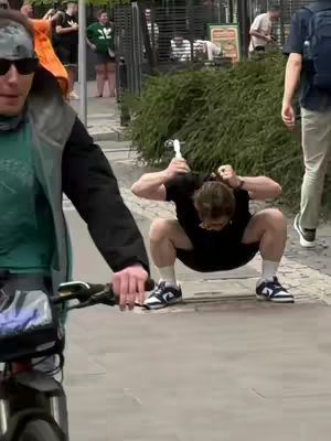 A street scene with a person squatting in the middle of the frame, near a person on a bike, and some people in the background. The person squatting is wearing a black shirt, shorts, and sneakers and is holding their head. The person on the bike is a man with a green jacket and sunglasses.