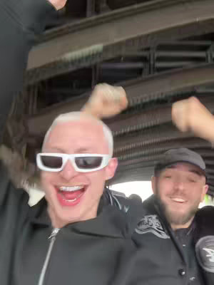 Two men are shown close up, smiling and cheering with their arms raised. The one on the left has short, bleached blonde hair, white sunglasses, and dental braces. The background is a blurred view of an underpass with metal structures.