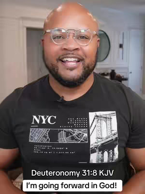 A medium shot features a Black man with glasses smiling at the camera. He has a neatly trimmed beard and is wearing a black t-shirt with a graphic print. The background is blurred, showing a glimpse of a white-walled room. Text at the bottom provides the scripture verse and message.