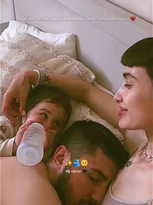 A family, including a mother, father, and young child, is snuggled together in bed. The child is drinking from a bottle. The mother is kissing the father.