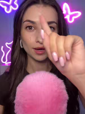 A woman with brown hair and fair skin is the focus, with a neon purple butterfly and lines in the background. She is holding a pink fluffy microphone in her right hand with her index finger pointed up.