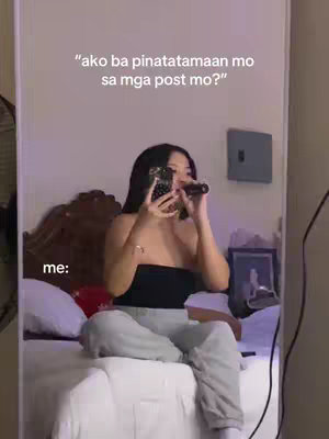 A person is sitting on a bed, holding up a phone in front of their face, as if taking a selfie. The background is a room with a mirror, headboard and side table. Text overlay on top reads a question in a foreign language.