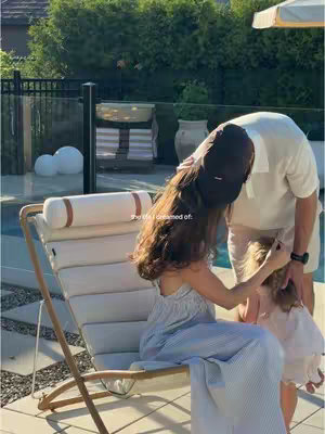 A sunny outdoor scene shows a family of three in a backyard setting. A woman is sitting and hugging her child, while a man is standing nearby. The sunlight creates a warm, inviting atmosphere.