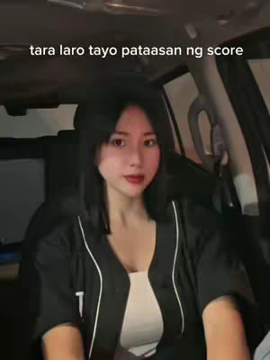 A woman with dark hair is seated in a car. She is wearing a black shirt over a white top and looking at the camera. Text overlaid reads, "tara laro tayo pataasan ng score". The background indicates a dimly lit environment.