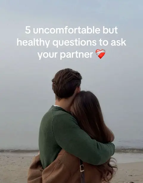 A couple embraces on a beach, seen from behind. The man in a green sweater and the woman with brown hair are the focus, with the ocean and sky forming a soft, blurred background.