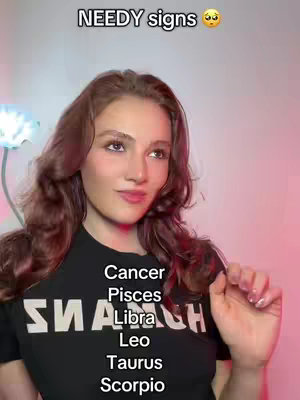 A young woman with long brown hair is centered in the frame, smiling slightly and glancing to the side. The background has soft pink and white lighting. A large black shirt with white text covers her torso. The foreground displays a list of zodiac signs.