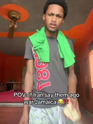 A young man stands indoors against an orange-toned wall with a patterned ceiling fan visible. He wears a gray t-shirt with a colorful graphic and a green towel around his neck. Text overlays are positioned at the bottom of the screen.