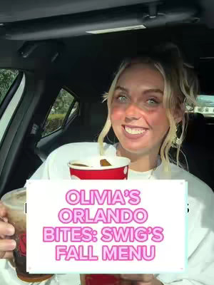 A woman in a car holds a cup of beverage and a sign with text on it. The text is in pink and white. The background is a car interior, suggesting a drive-through or to-go experience.