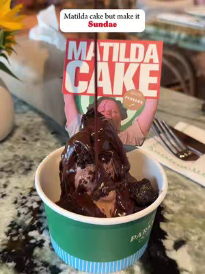 A sundae in a green cup with chocolate sauce being poured over it, along with pieces of cake. A paper sign that says 'Matilda Cake' is stuck in the sundae.