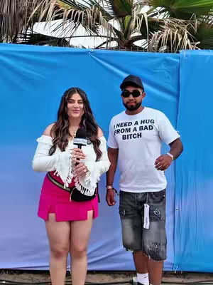 A photo with a smiling woman with long, wavy, dark hair wearing a white, off-the-shoulder blouse and a short, bright pink skirt standing with a man in a white t-shirt and sunglasses. They are both facing the camera, and the woman is holding a black microphone. A blue background, likely a backdrop, is behind them with some palm trees visible.