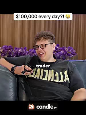 A young man is seated on a dark blue couch in front of a wooden wall covered in purple flowers. He is wearing black glasses and a black t-shirt with white capital letters. He smiles and holds a microphone, while text overlays read '$100,000 every day?! π€£' and 'trader'.