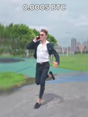 A man in a suit runs with a phone in his ear on a pathway, with blurred background of green trees and a city skyline. The text overlay in white reads '0.0005 BTC'.