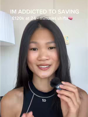 A young woman with long dark hair is centered in a well-lit shot, looking directly at the camera with a slight smile. She's wearing a black top, and a small microphone is visible near her mouth. The background is softly blurred, with a lamp and wall visible.