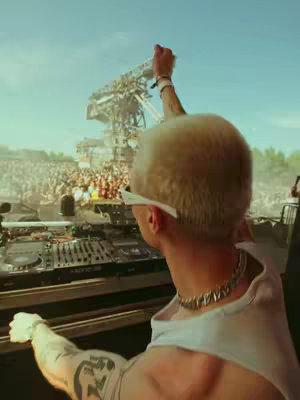 The image features a person, viewed from the back, performing at what appears to be an outdoor music festival or concert. The person is wearing sunglasses and a white top and has short, light-colored hair. They are facing towards a large crowd, with one arm raised. In the background, there's a large stage setup and a multitude of people. The sky is clear with daylight. DJ equipment is visible in the foreground.