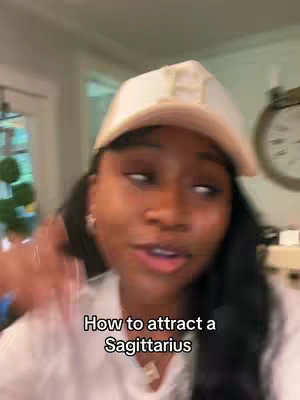 A woman with long dark hair, wearing a white baseball cap and a white shirt, is looking at the camera and talking. Text on the image reads: How to attract a Sagittarius. The background is slightly blurred, showing interior details.