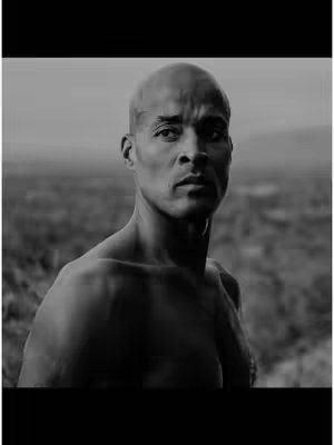 A black and white image featuring a bald man in the center, looking directly at the camera with a serious expression. He's partially shirtless, showing his upper body and shoulders. The background is blurred, suggesting an outdoor setting with a subtle landscape. The image has a focus on the man's face, with strong lighting and shadows.