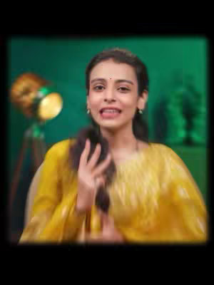 A woman with dark hair is sitting and talking. She is wearing a yellow top and there is a dim green background as well as a spotlight. The image is slightly blurry, and has a vignette effect.