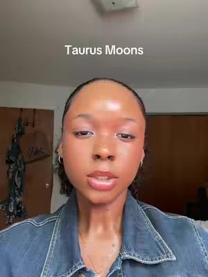 A close-up shot features a woman looking directly at the camera with a neutral expression. The background is a room with a door and a light fixture. The woman has clear skin and is wearing a denim jacket. The text 'Taurus Moons' is overlaid at the top of the frame in white.